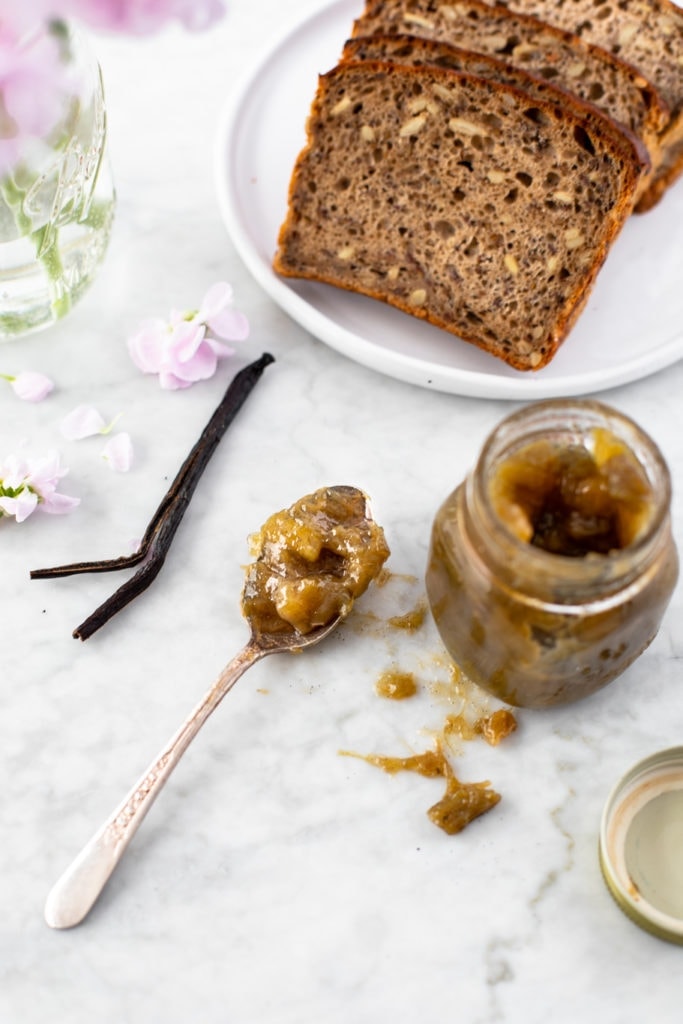 Spoon with rhubarb vanilla jam, next to an open jar. Vanilla bean, slices of bread and flowers
