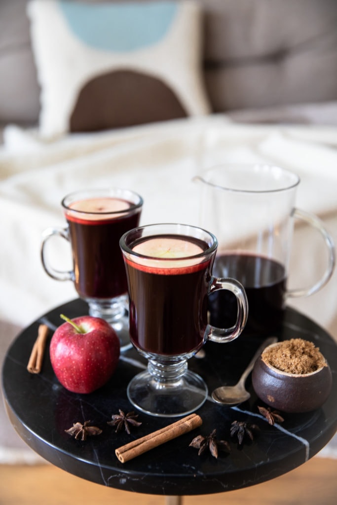 Glass mugs with red mulled wine, arranged on a coffee table with red apple, cinnamon sticks, star anise, brown sugar, and teaspoon
