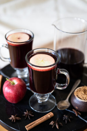 Glass mugs with red mulled wine, arranged on a coffee table with red apple, cinnamon sticks, star anise, brown sugar, and teaspoon
