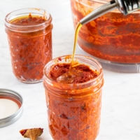 Photo of jars filled with spicy chili paste harissa, and being topped with a thin layer of olive oil to preserve it | from verygoodcook.com